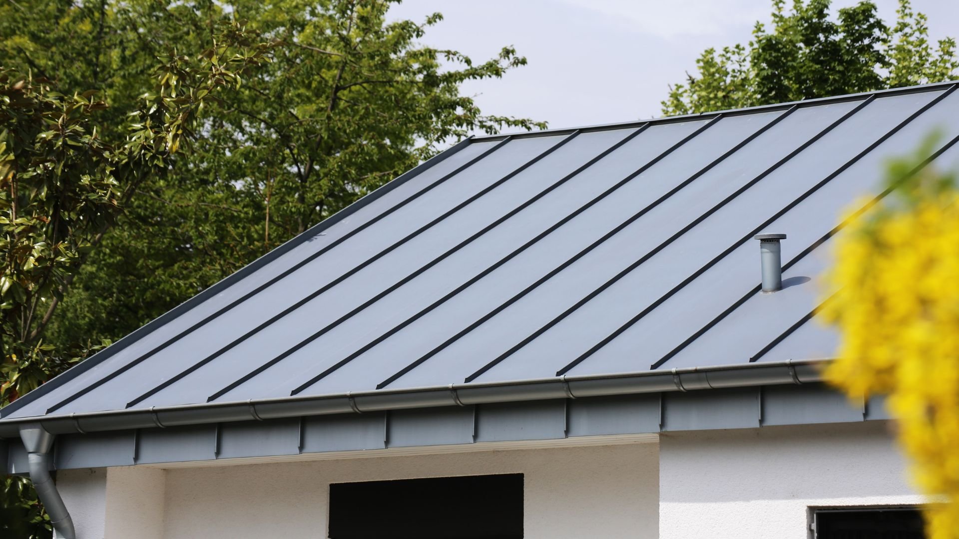 Gray metal roof with vertical seams on white building surrounded by trees