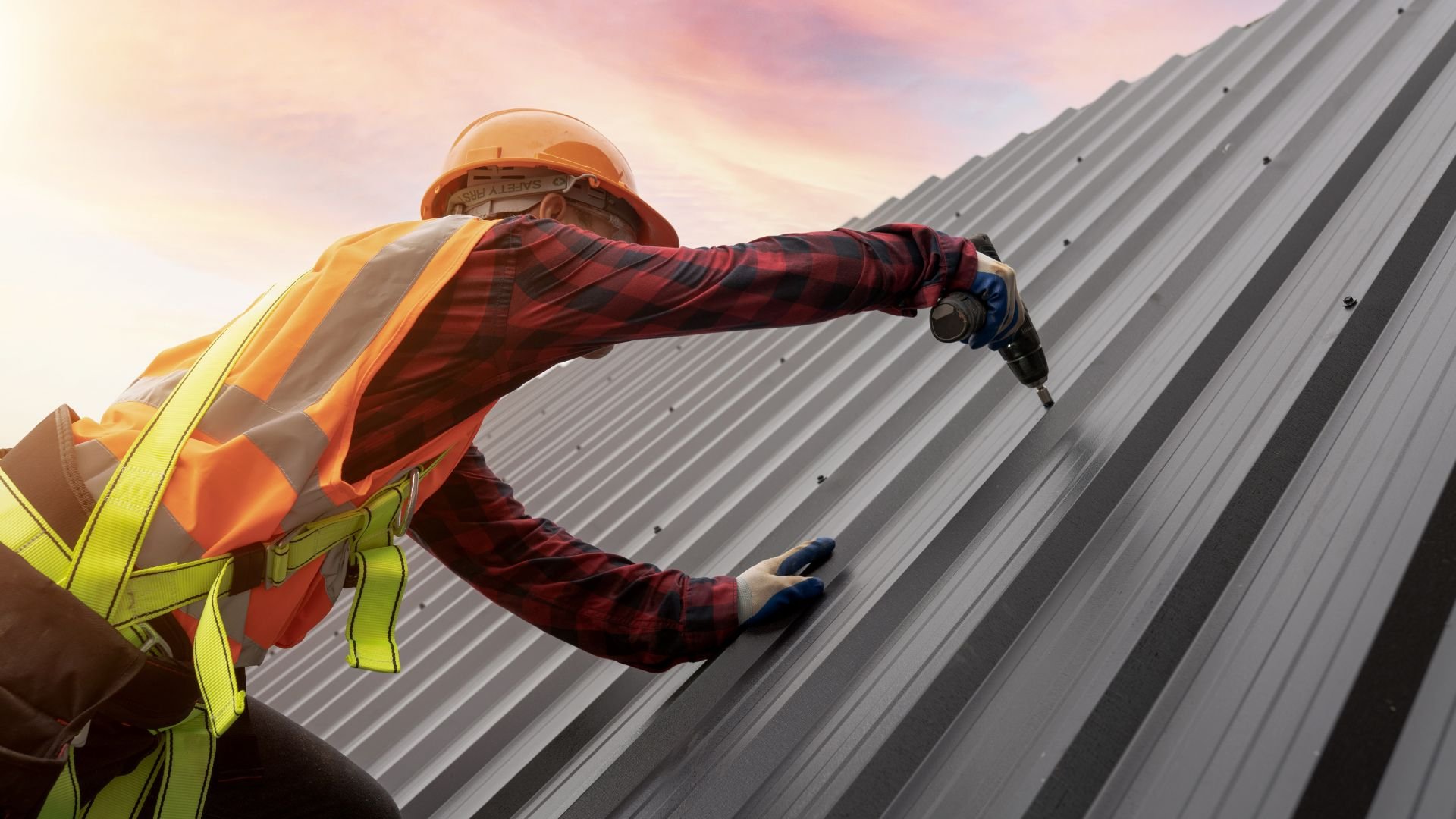 Construction worker installing metal roofing with drill at sunset