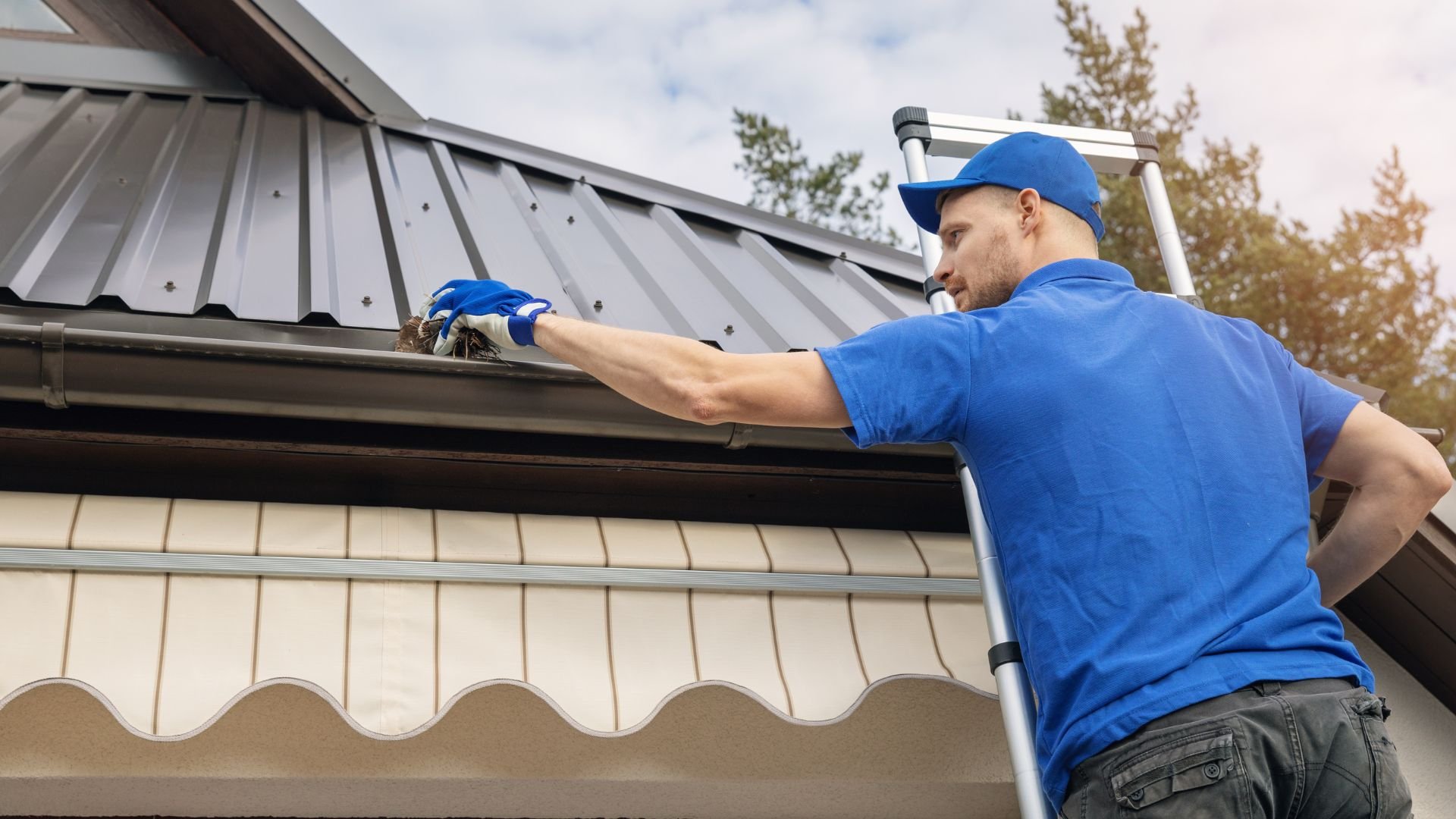 Worker in blue cleaning gutters on metal roof using ladder
